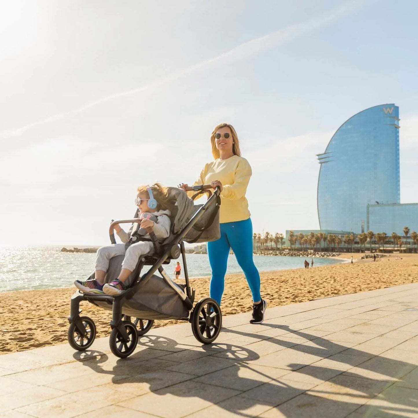 Woman pushing a stroller with a child on a beach, with a modern building in the background.
