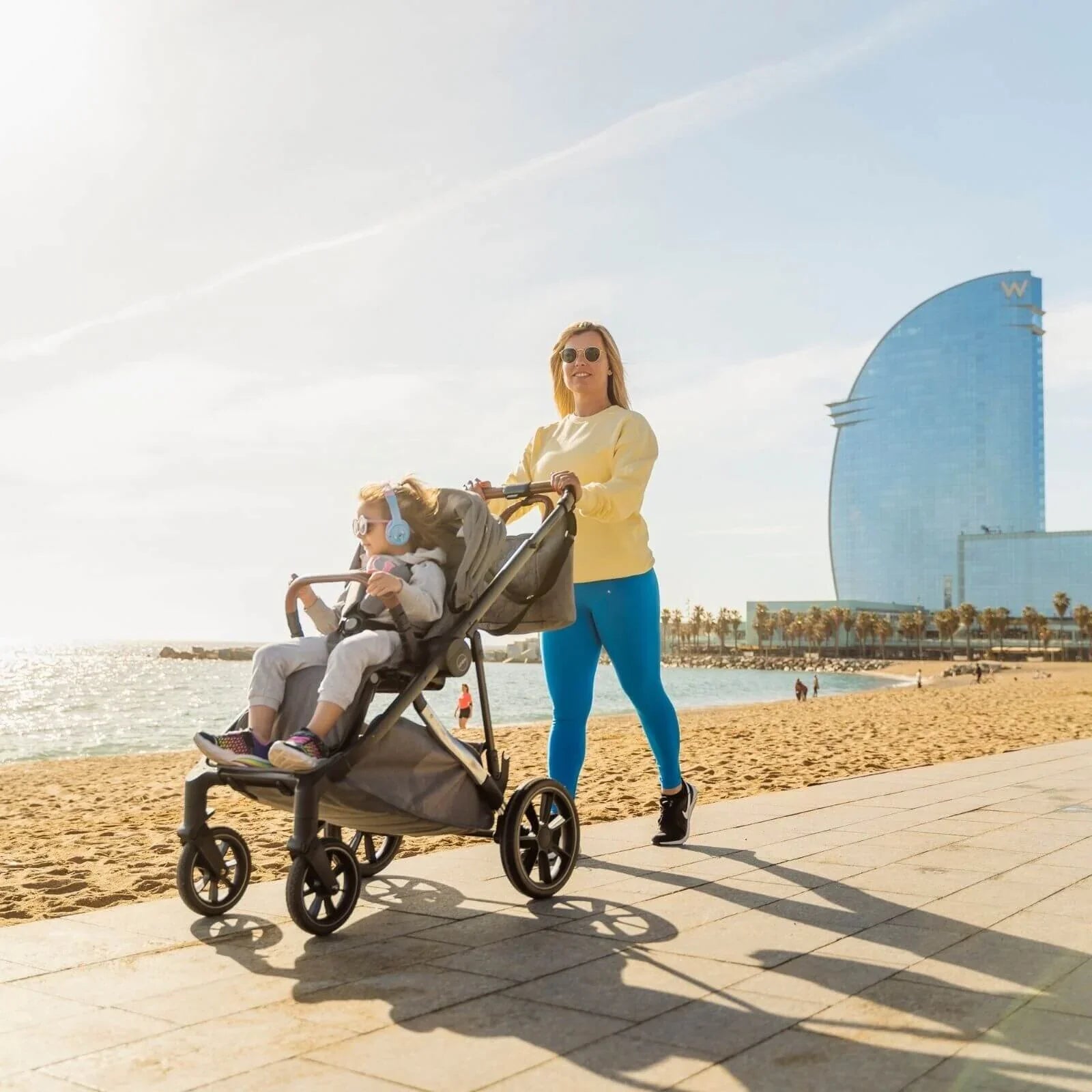 Woman pushing a stroller with a child on a beach, with a modern building in the background.